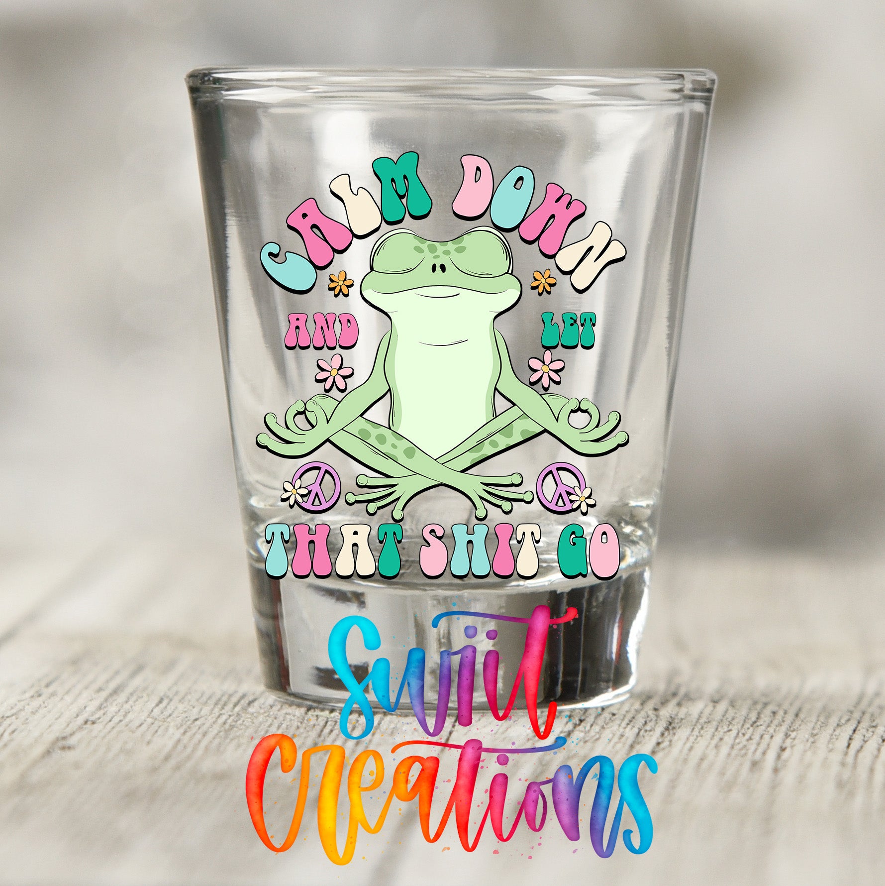 A shot glass with a colorful design featuring a frog and the text "Calm down and let go that shit go".