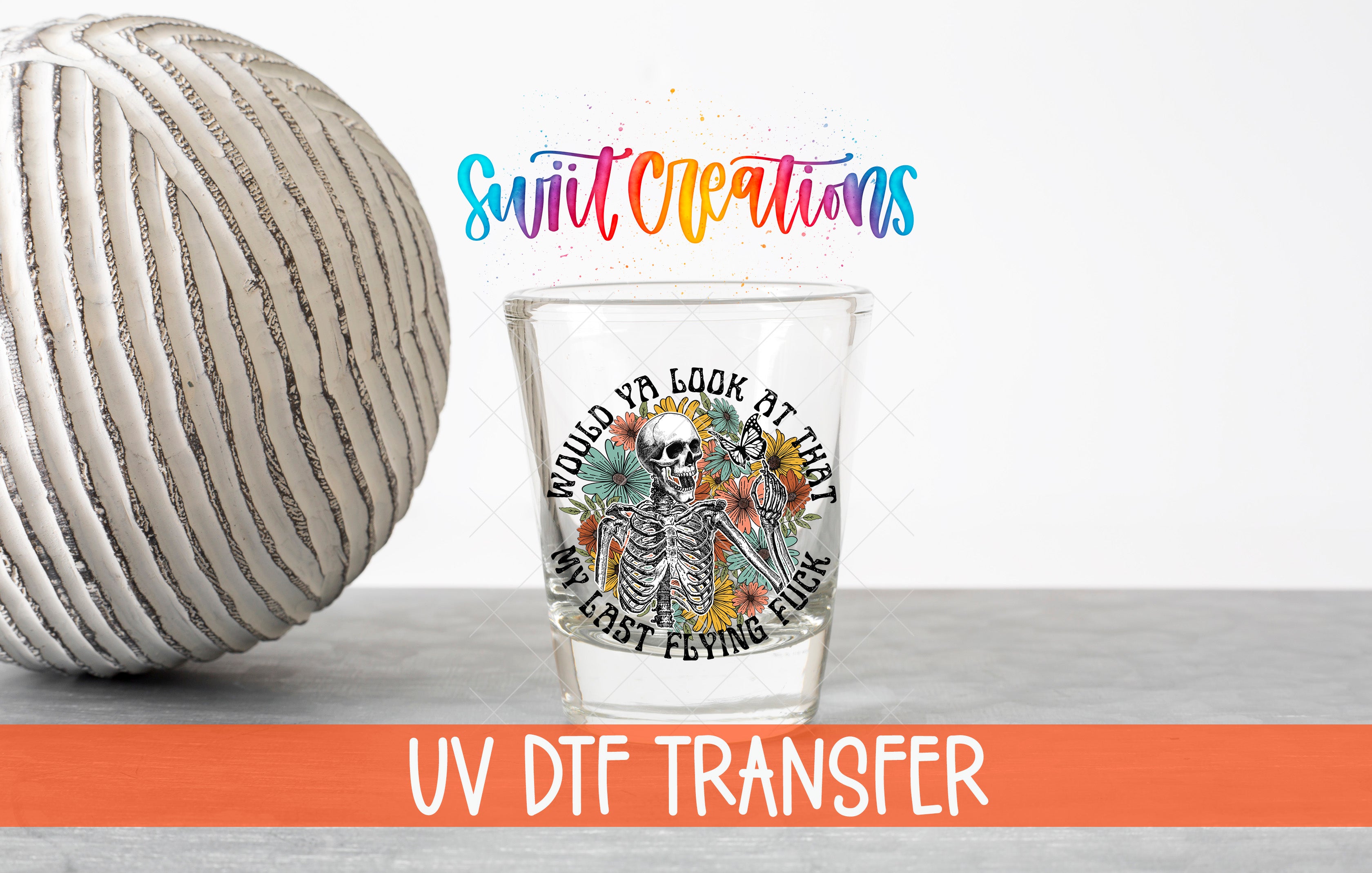 a glass with a skeleton design and text, placed next to a white ball on a gray surface.