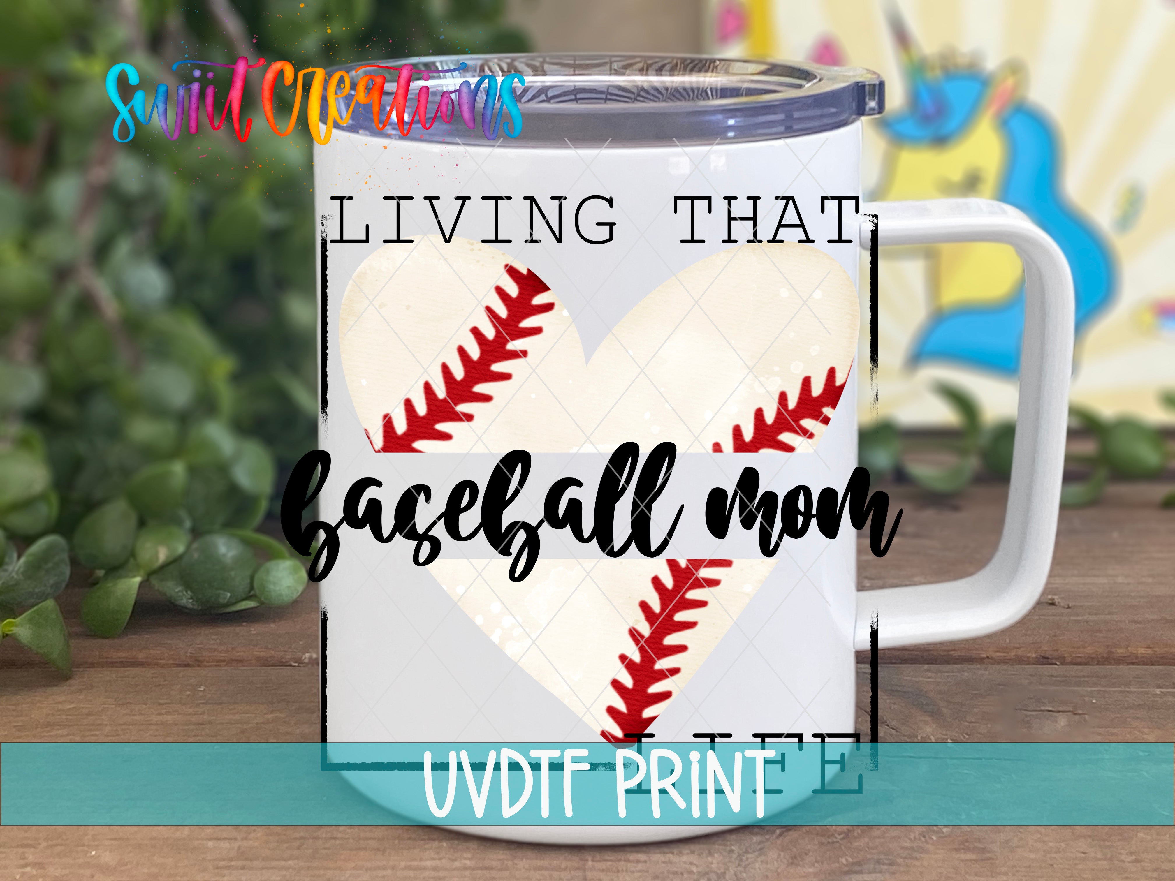 A white mug with a baseball design and the text "LIVING THAT BASEBALL MOM" printed on it, placed on a wooden surface with green leaves in the background.