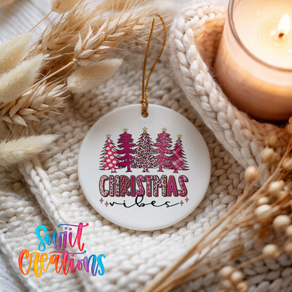 A white Christmas ornament with a design of Christmas trees and the text "Christmas vibes" hangs on a white knit blanket. A lit candle is also present in the scene.