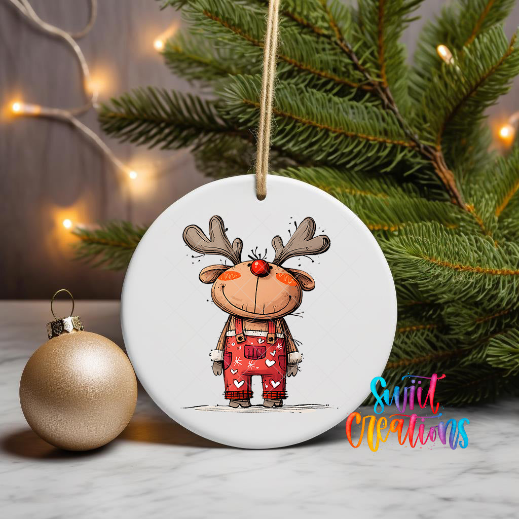 A white ceramic ornament featuring a cartoon reindeer wearing overalls and antlers, hanging on a string next to a gold ornament and a Christmas tree.