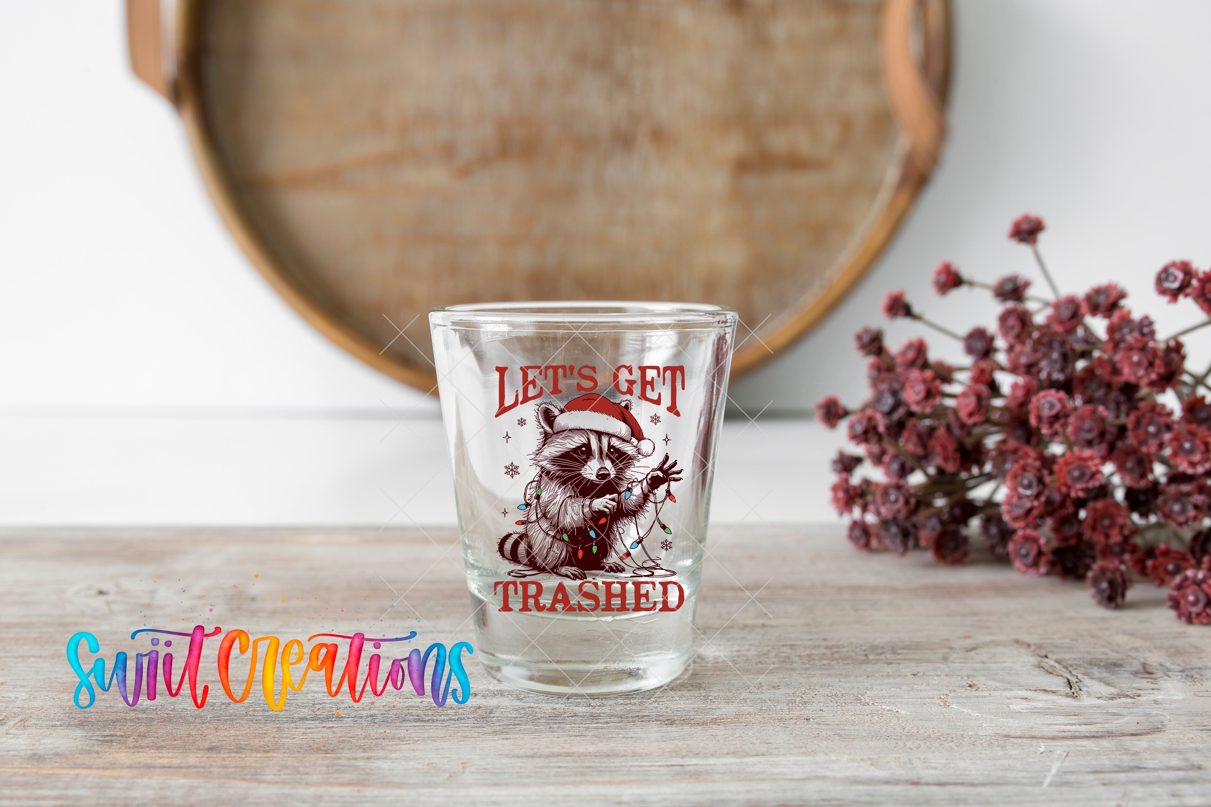 A shot glass with a raccoon wearing a Santa hat and the text "Let's Get Trashed" printed on it, placed on a wooden surface next to a bunch of red flowers.