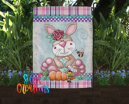 A garden flag featuring a cartoon rabbit with a flower on its head, surrounded by flowers and plants.