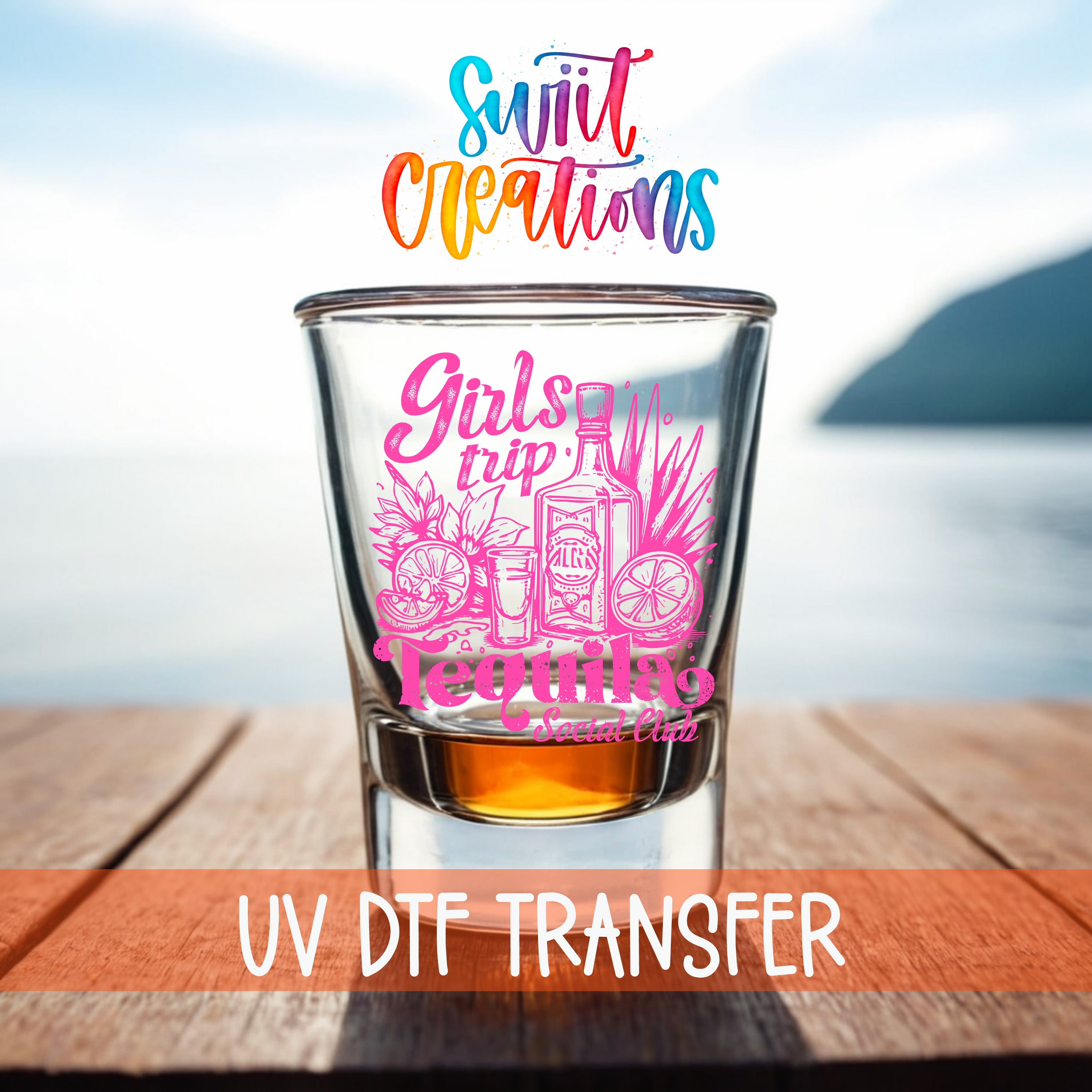 A glass of tequila with a colorful design on it, placed on a wooden surface with a body of water in the background.