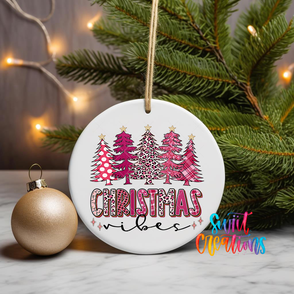 A white Christmas ornament with a pink and leopard print design, featuring the text "Christmas vibes" and surrounded by Christmas trees. The ornament is hanging from a string, and there is a gold ornament next to it on a table.