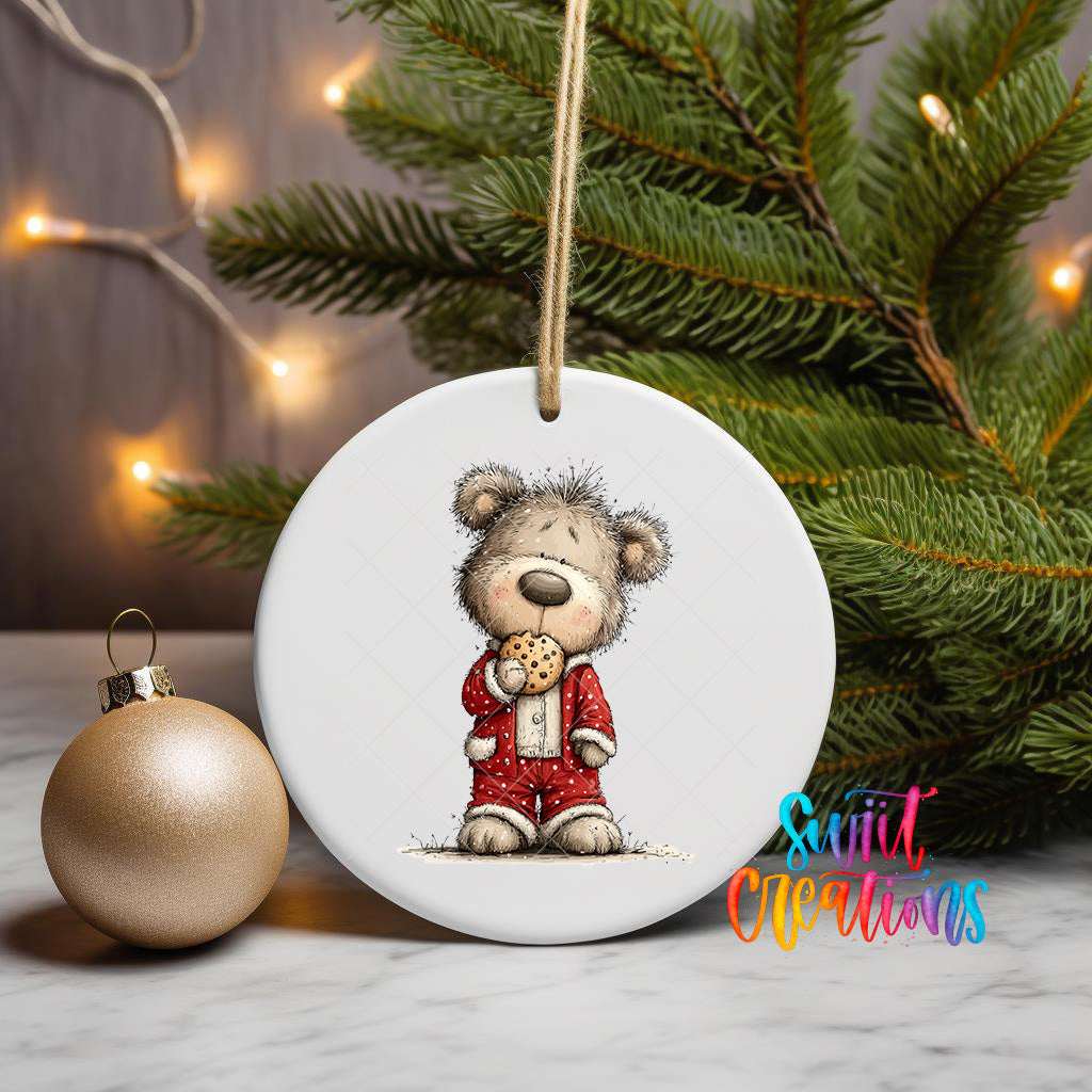 A white ceramic ornament featuring a teddy bear wearing a red outfit and holding a cookie, placed on a marble surface next to a gold ornament and a Christmas tree with twinkling lights.