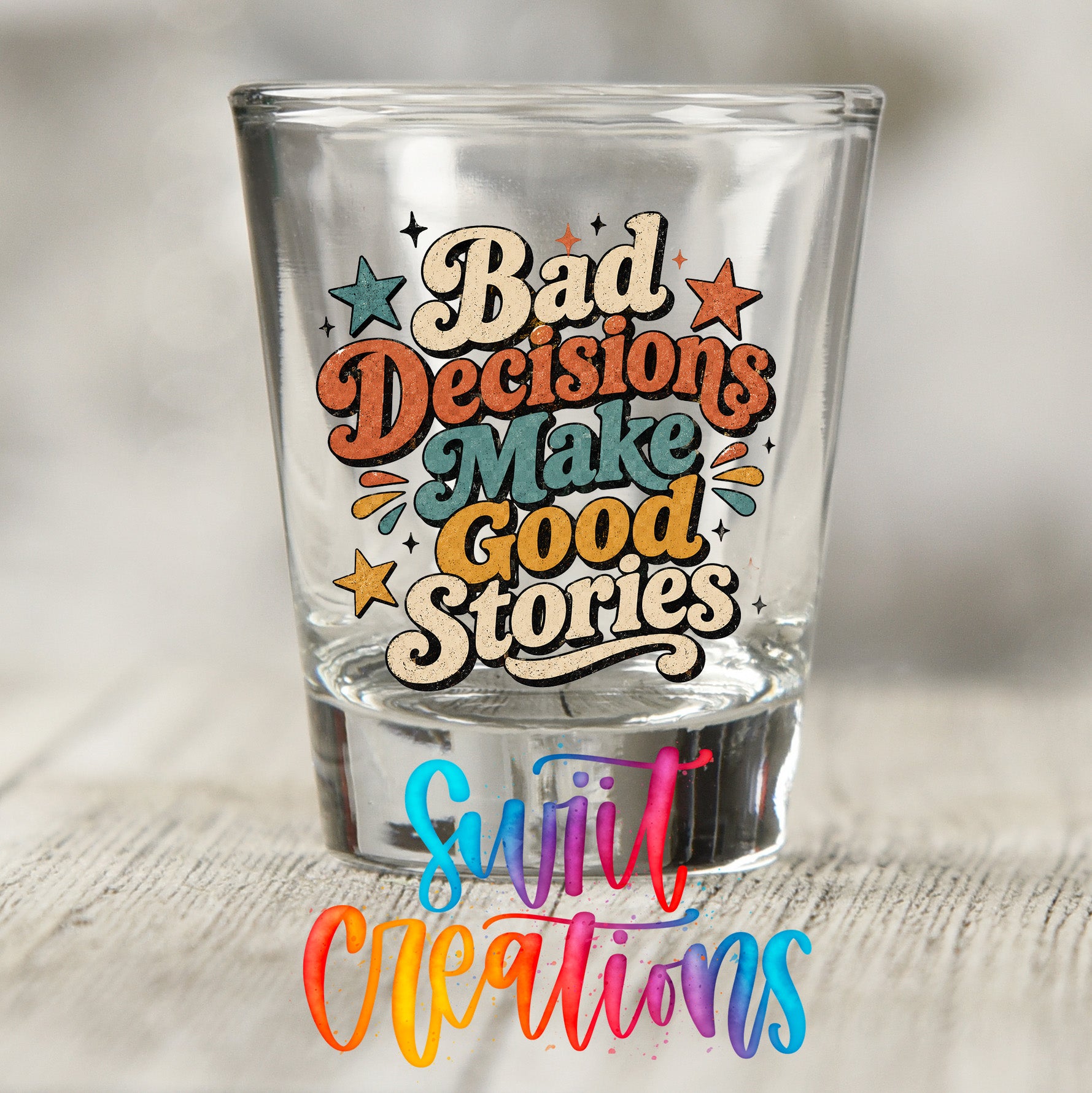 A shot glass with a colorful and playful design featuring the text "Bad Decisions Make Good Stories" surrounded by stars and other decorative elements.
