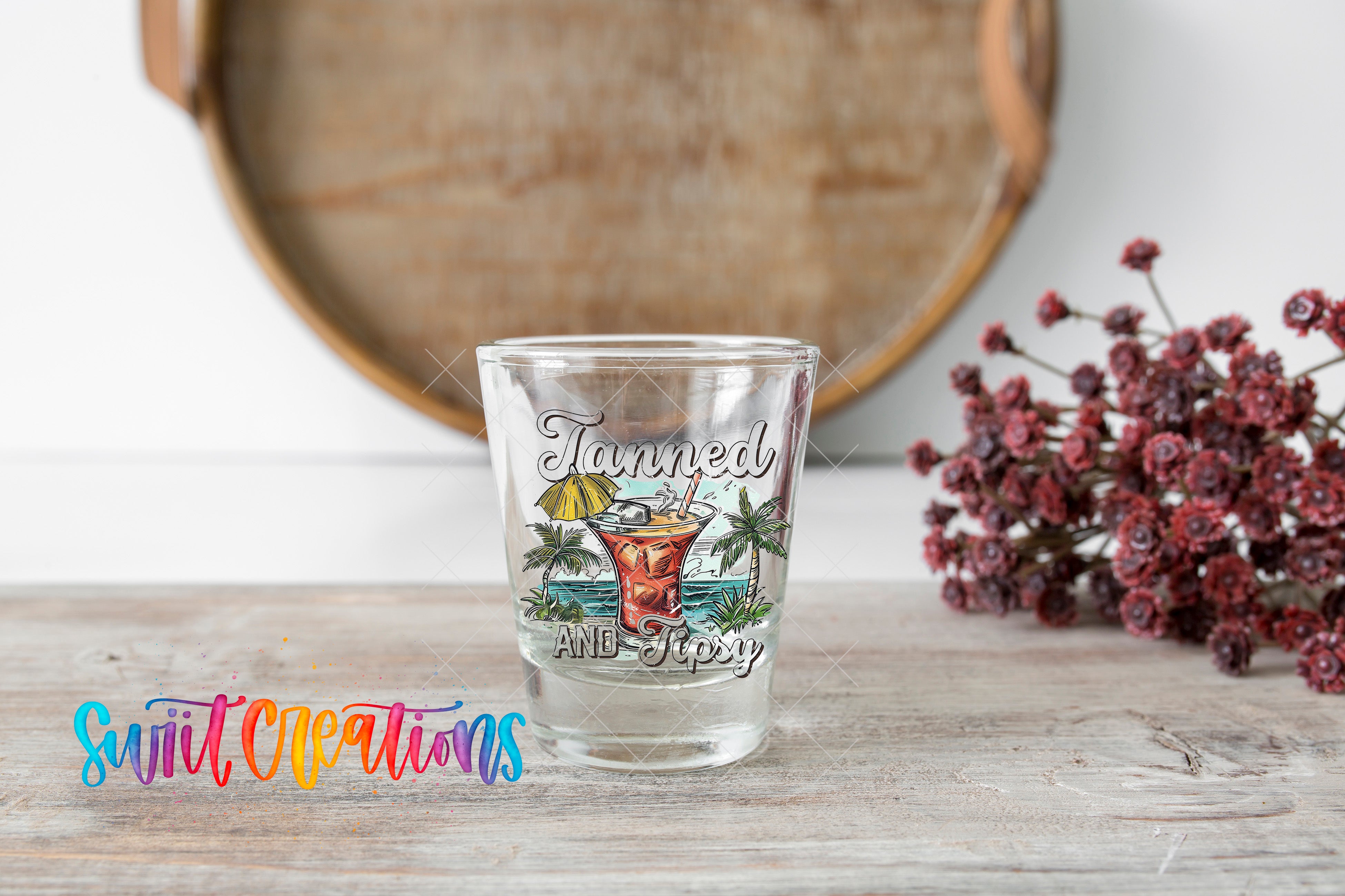 A shot glass with a tropical drink design and the text "Tanned and sticky" is placed on a wooden surface, with a bouquet of flowers and a wooden tray in the background.