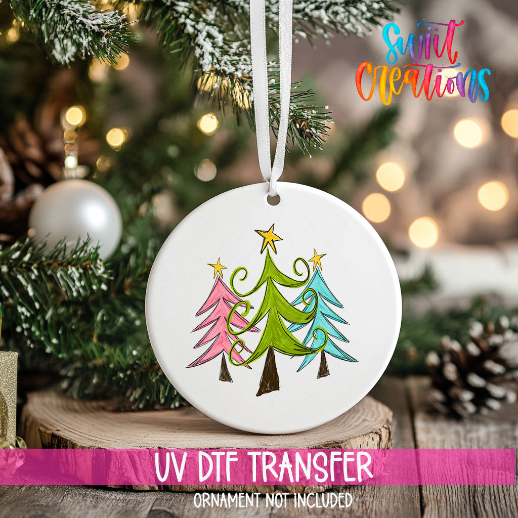 A white ceramic ornament with a colorful illustration of three Christmas trees hangs on a tree branch, with a blurred background of twinkling lights.