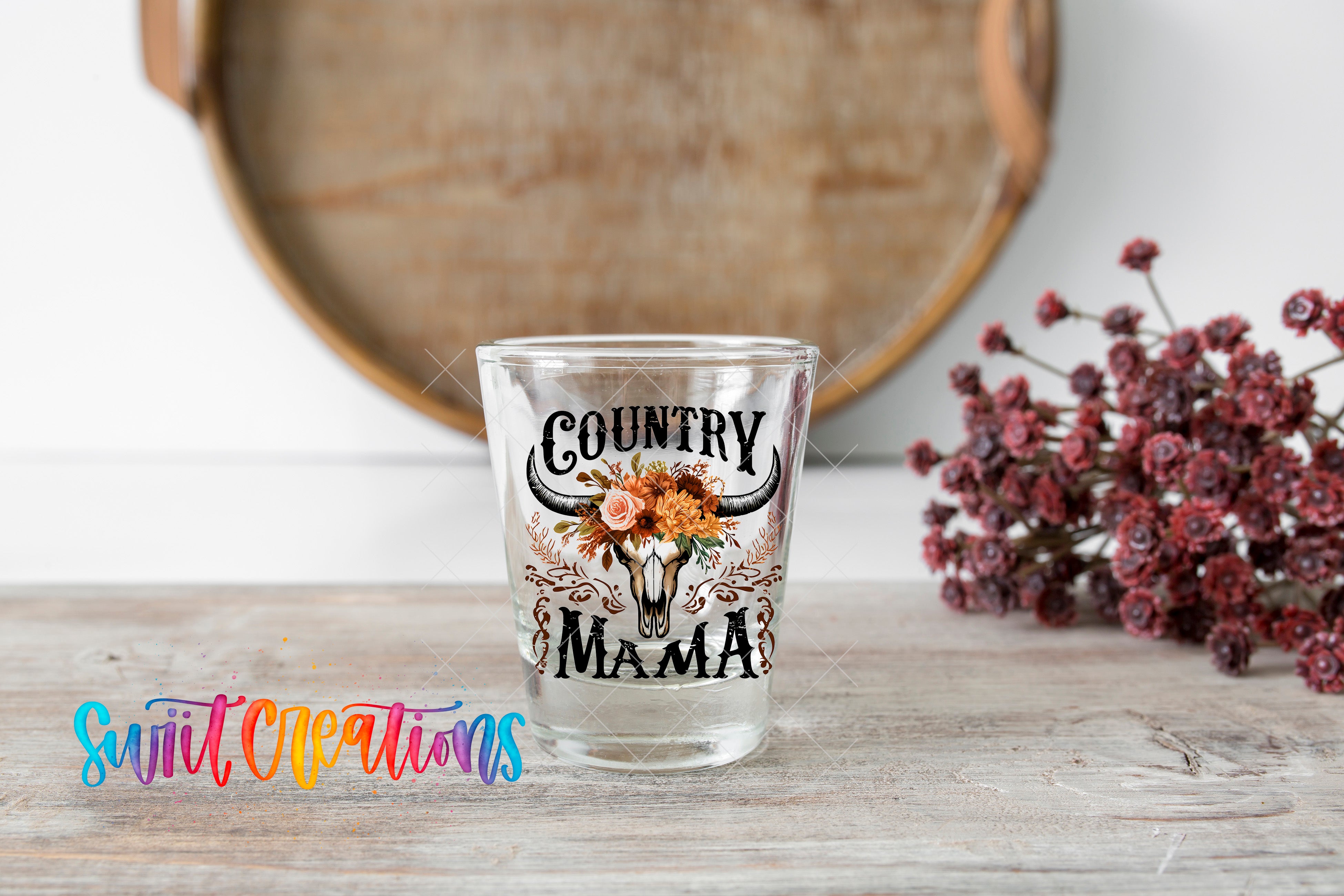 A glass shot glass with the text "COUNTRY MAMA" printed on it, placed on a wooden surface next to a bunch of red flowers.