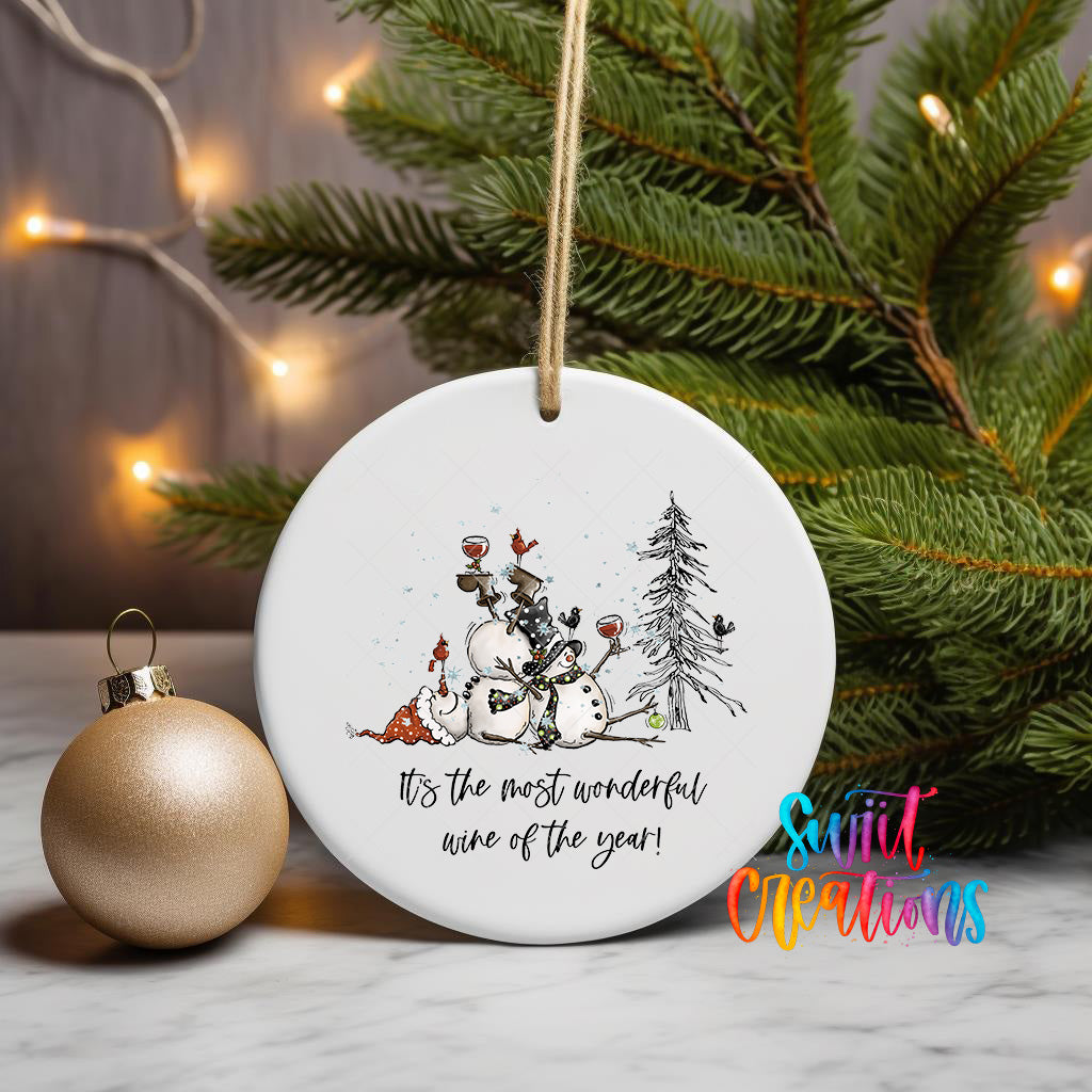 A white ceramic ornament with a snowman and Christmas tree design, along with a text that says "It's the most wonderful wine of the year!" is displayed on a table, with a gold ornament and a Christmas tree in the background.