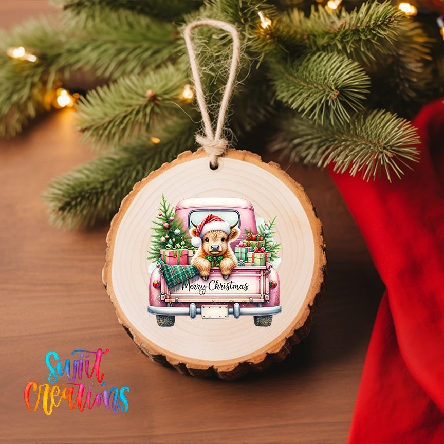 A wooden ornament hanging on a Christmas tree, featuring a cow wearing a Santa hat and carrying presents in the back of a pink truck.