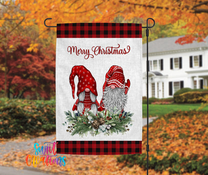 A banner with a Christmas-themed design featuring two gnomes wearing red hats and scarves, surrounded by a wreath of pine cones and other holiday decorations. The banner is hanging outside a house with autumn foliage in the background.