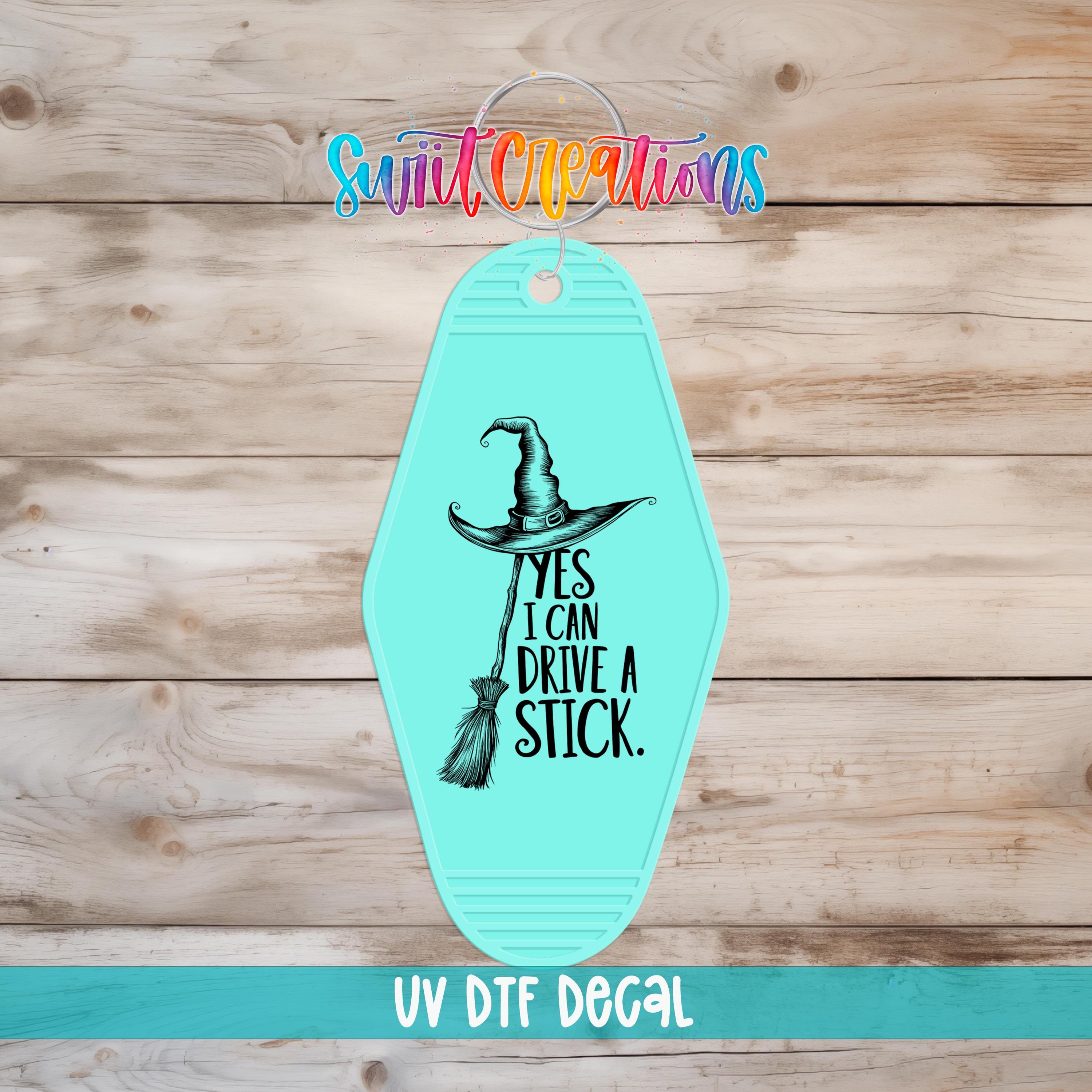 A turquoise-colored keychain with a witch's hat and the text "YES I CAN DRIVE A STICK" printed on it, along with the text "UV Dtf Decal" at the bottom.