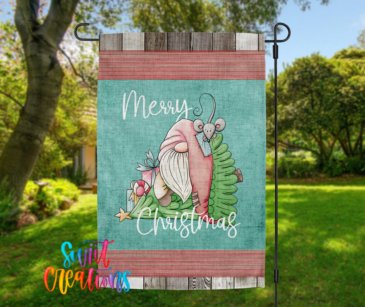 A colorful garden flag with a gnome and Christmas decorations, including a tree and presents, hanging in a grassy area with trees in the background.