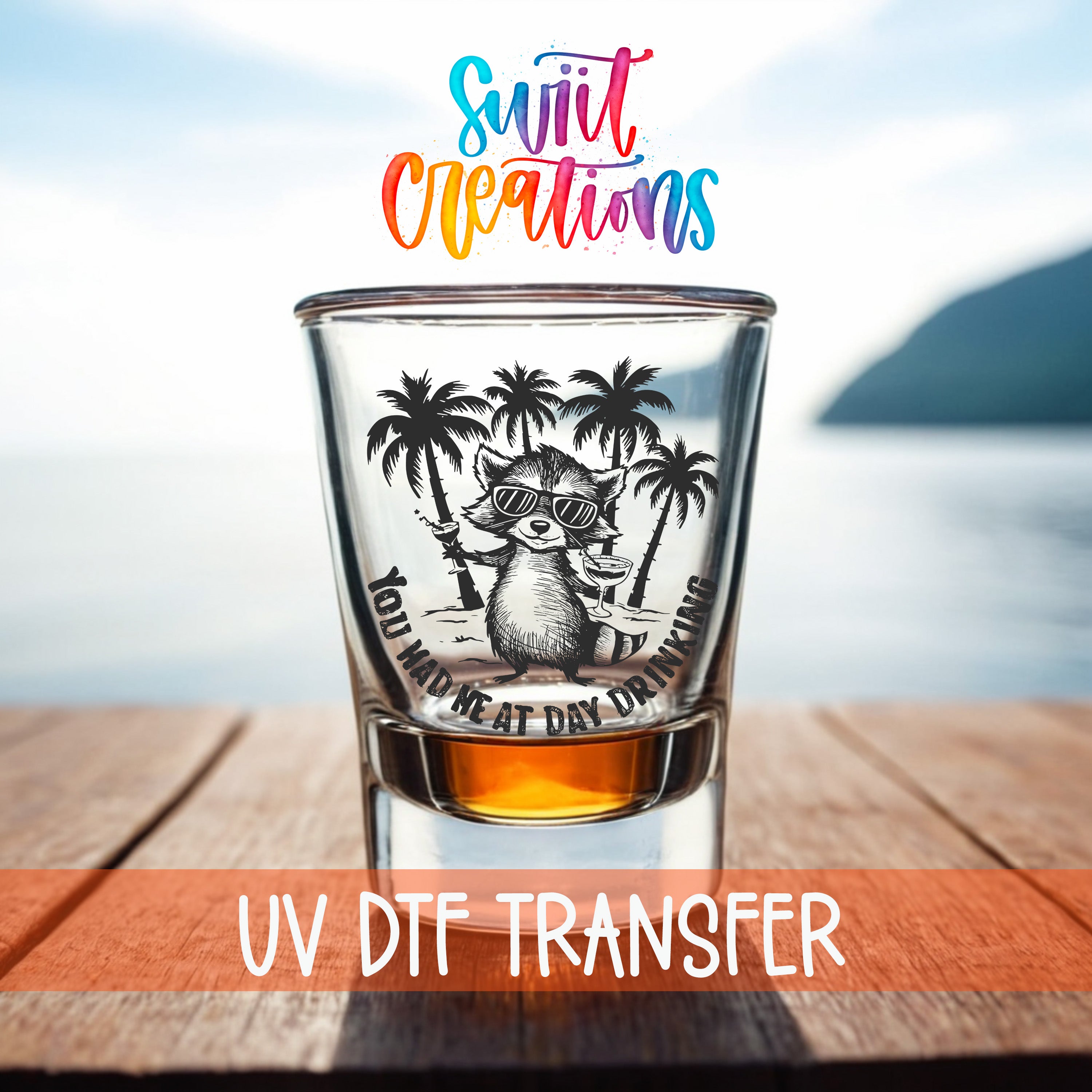 A shot glass with a cartoon raccoon and palm trees printed on it, placed on a wooden surface with a body of water in the background.