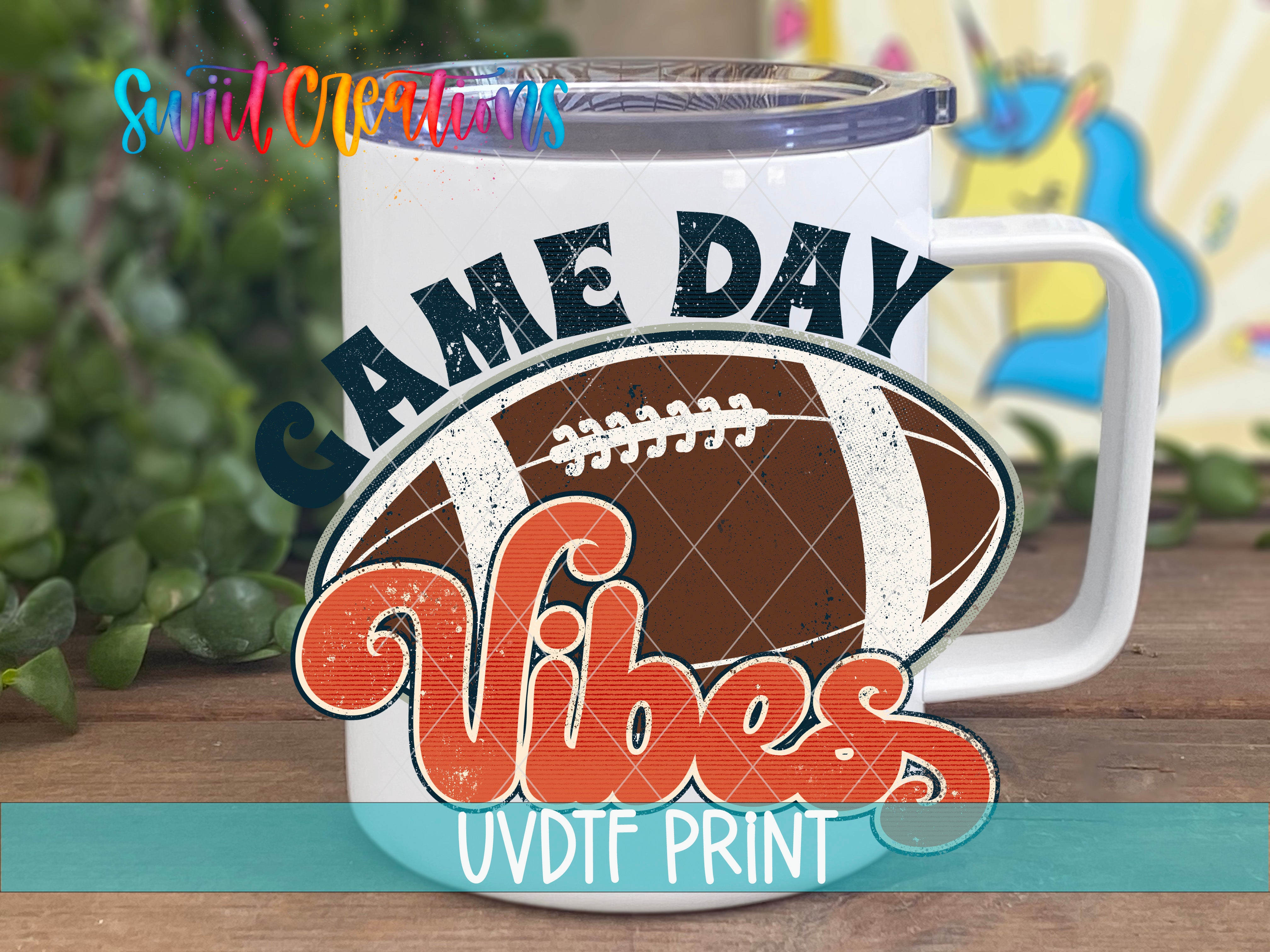A white mug with a football graphic and the text "GAME DAY VIBES" printed on it, placed on a wooden surface with greenery in the background.