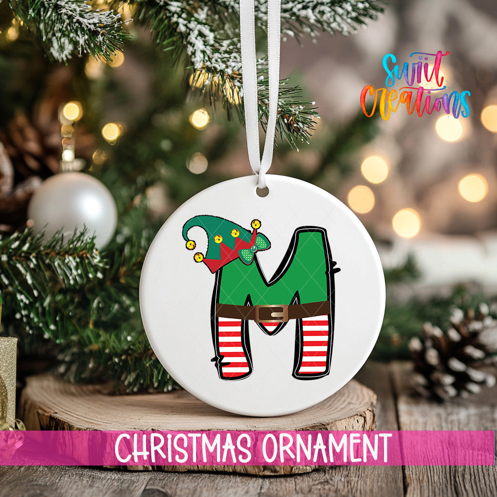 A Christmas ornament in the shape of the letter "M" with a green elf hat on top, hanging on a tree.