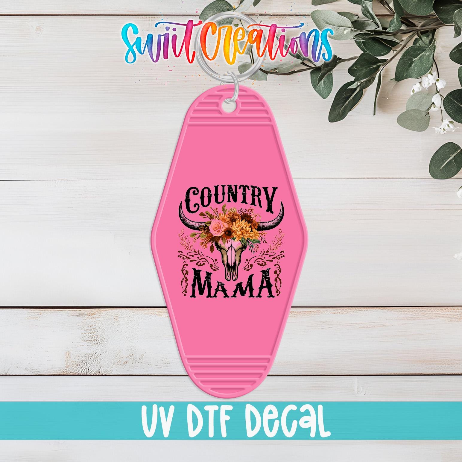 A pink keychain with the text "COUNTRY MAMA" and a floral design, placed on a wooden surface with a plant in the background.