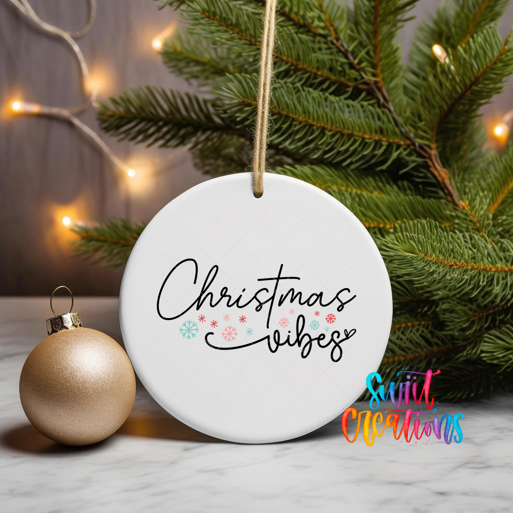 A white Christmas ornament with the text "Christmas vibes" written on it, hanging from a string. The ornament is placed next to a gold ornament and a pine tree branch with twinkling lights.