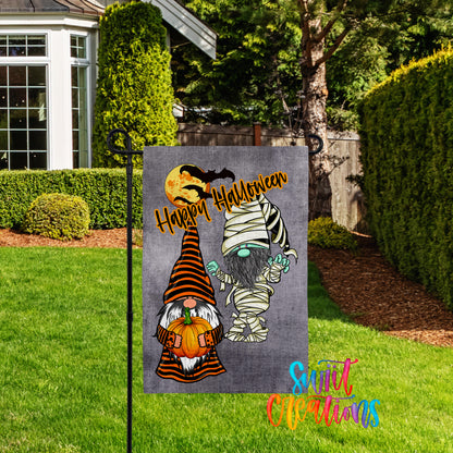 A Halloween-themed garden sign featuring two gnomes, one with a pumpkin and the other with a skeleton mask, standing in a yard with a house and trees in the background.