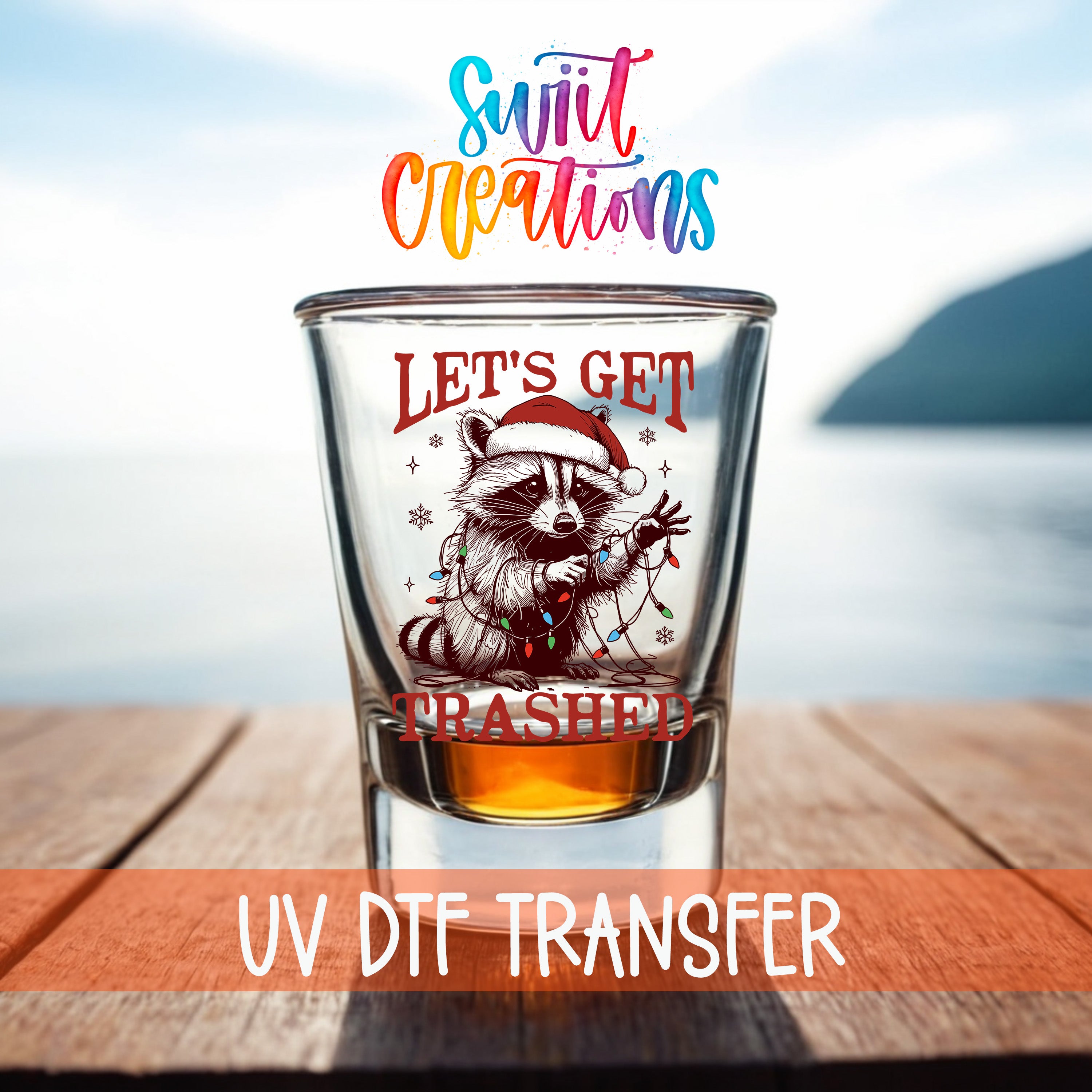 A shot glass with a raccoon wearing a Santa hat and the text "Let's get trampled" printed on it, placed on a wooden surface with a body of water in the background.