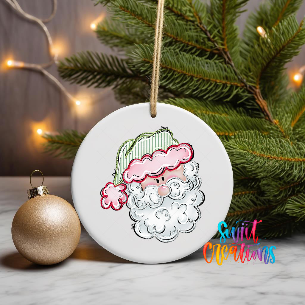 A white ceramic ornament with a Santa Claus face design is displayed on a marble surface, accompanied by a gold ornament and a Christmas tree with twinkling lights in the background.