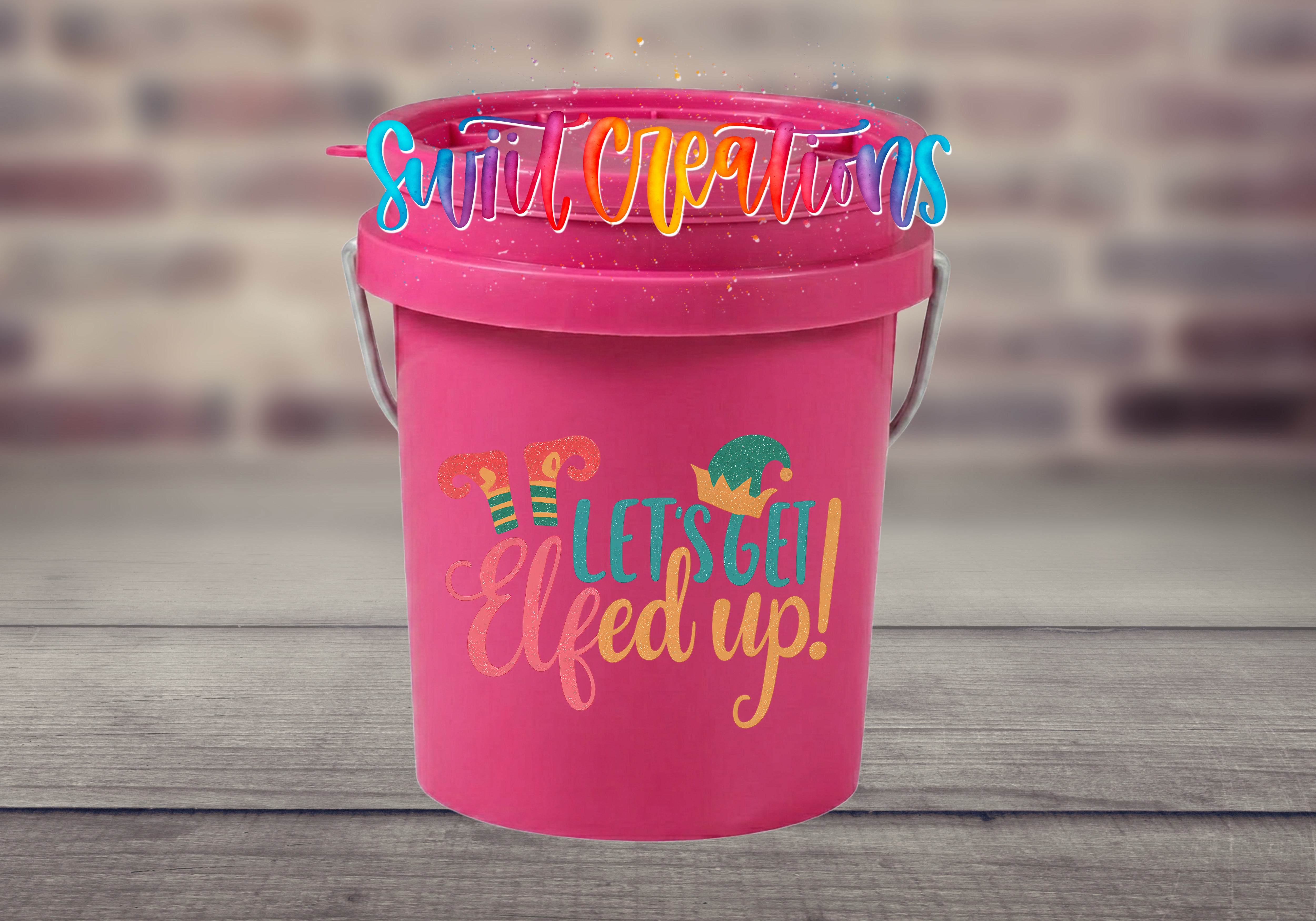 A pink bucket with a whimsical design and text on it, placed on a wooden surface against a blurred brick wall background.
