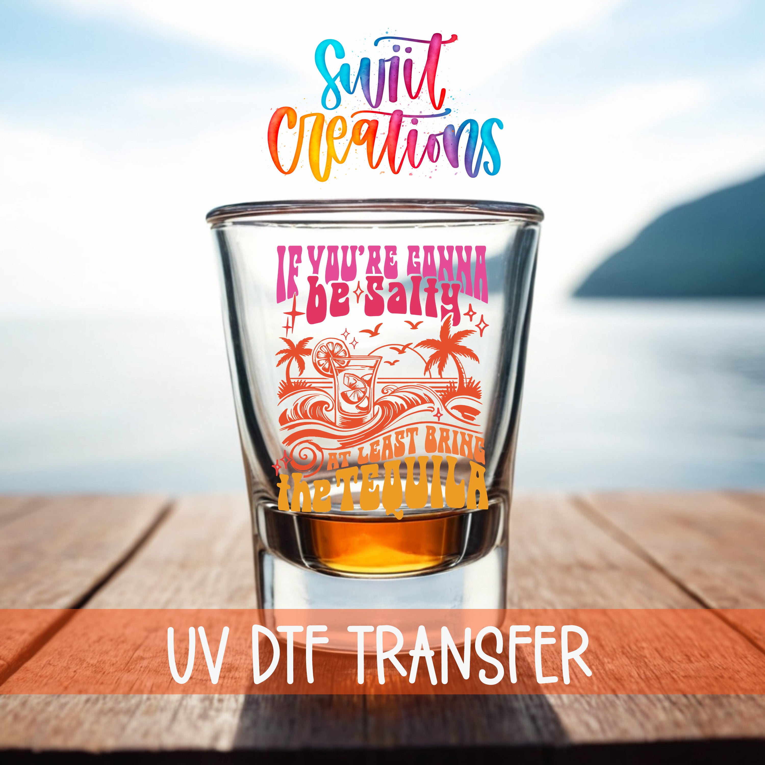 a shot glass with a colorful design and text, placed on a wooden surface with a body of water in the background.