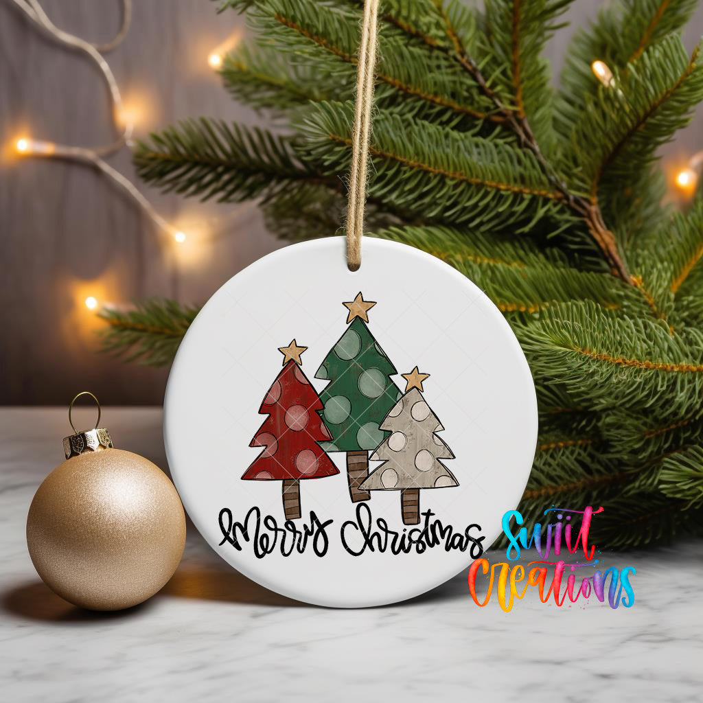 A white Christmas ornament with a design of three trees, one red, one green, and one white, is hanging from a string. The ornament is placed next to a gold ornament and a pine tree branch with twinkling lights.