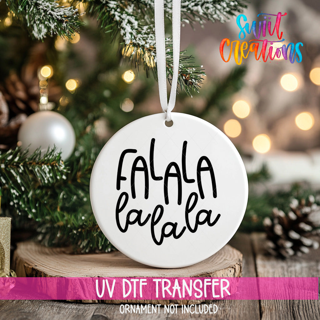 A white ornament with the text "FALA LA LA" hanging on a Christmas tree, with a blurred background of pine branches and twinkling lights.
