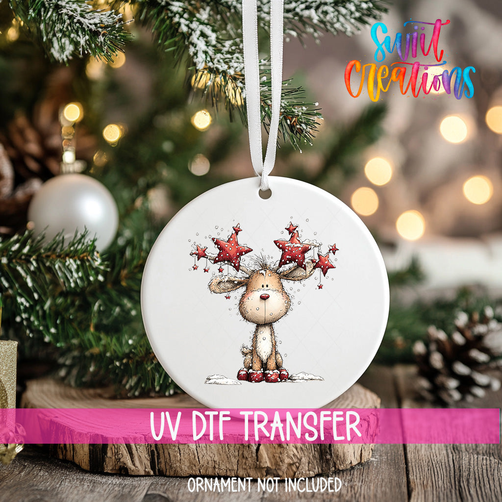 A white ornament with a cartoon reindeer and star decorations hangs on a Christmas tree, with a pink ribbon around it.