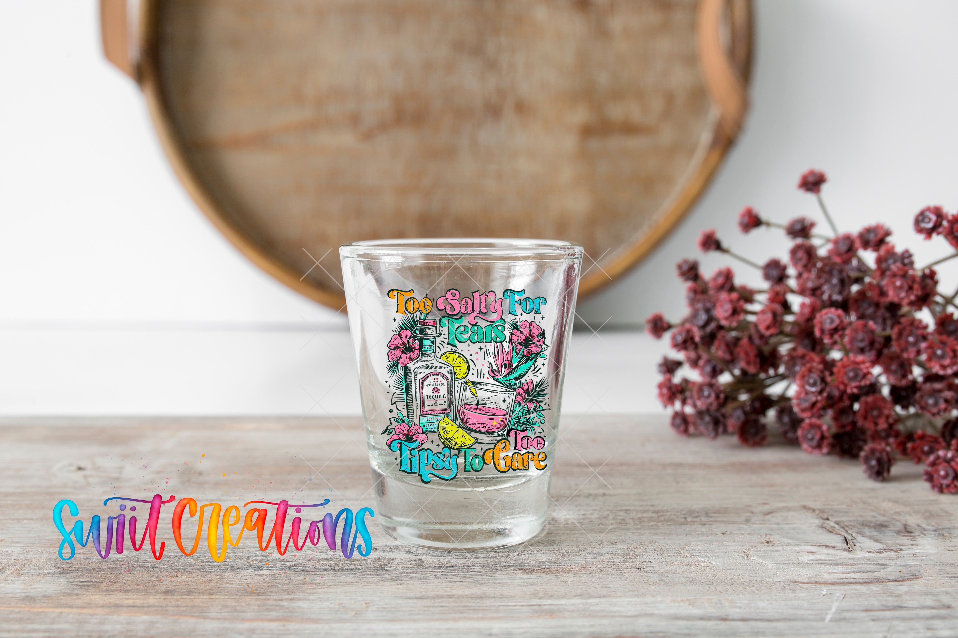 A shot glass with a colorful, whimsical design sits on a wooden surface, with a bouquet of flowers and a wooden tray in the background.