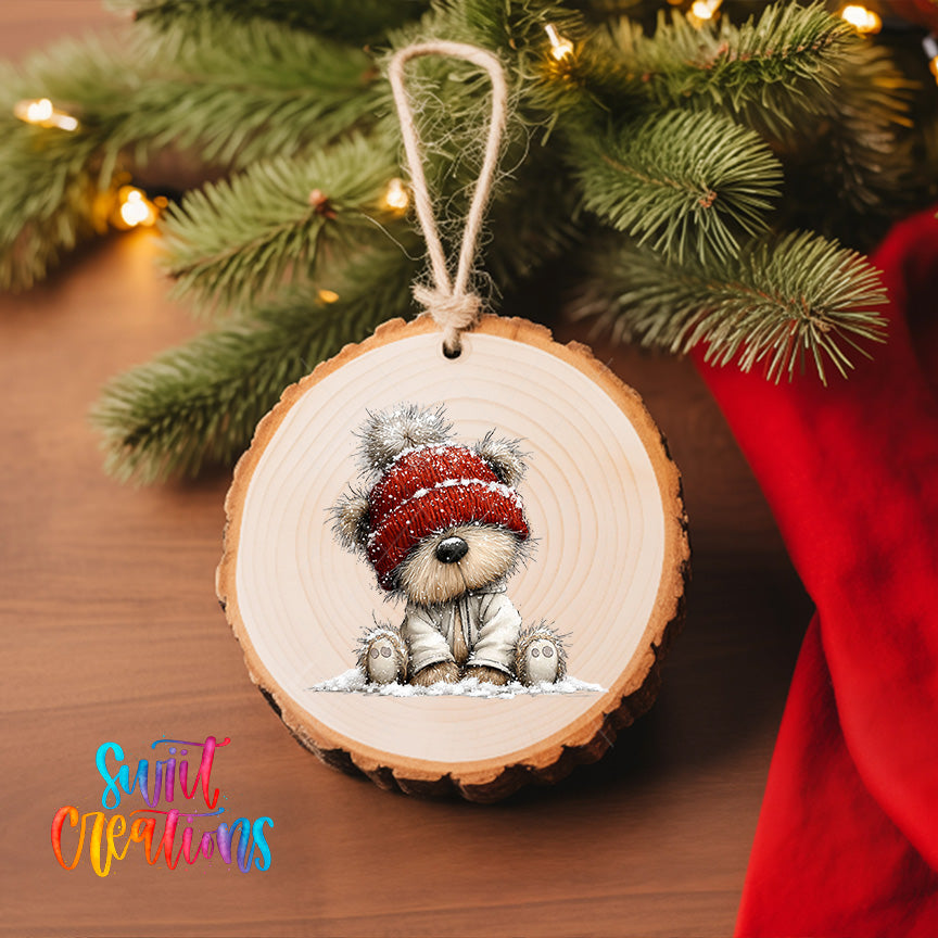 A wooden ornament featuring a teddy bear wearing a red hat, sitting in the snow, is hanging on a Christmas tree.
