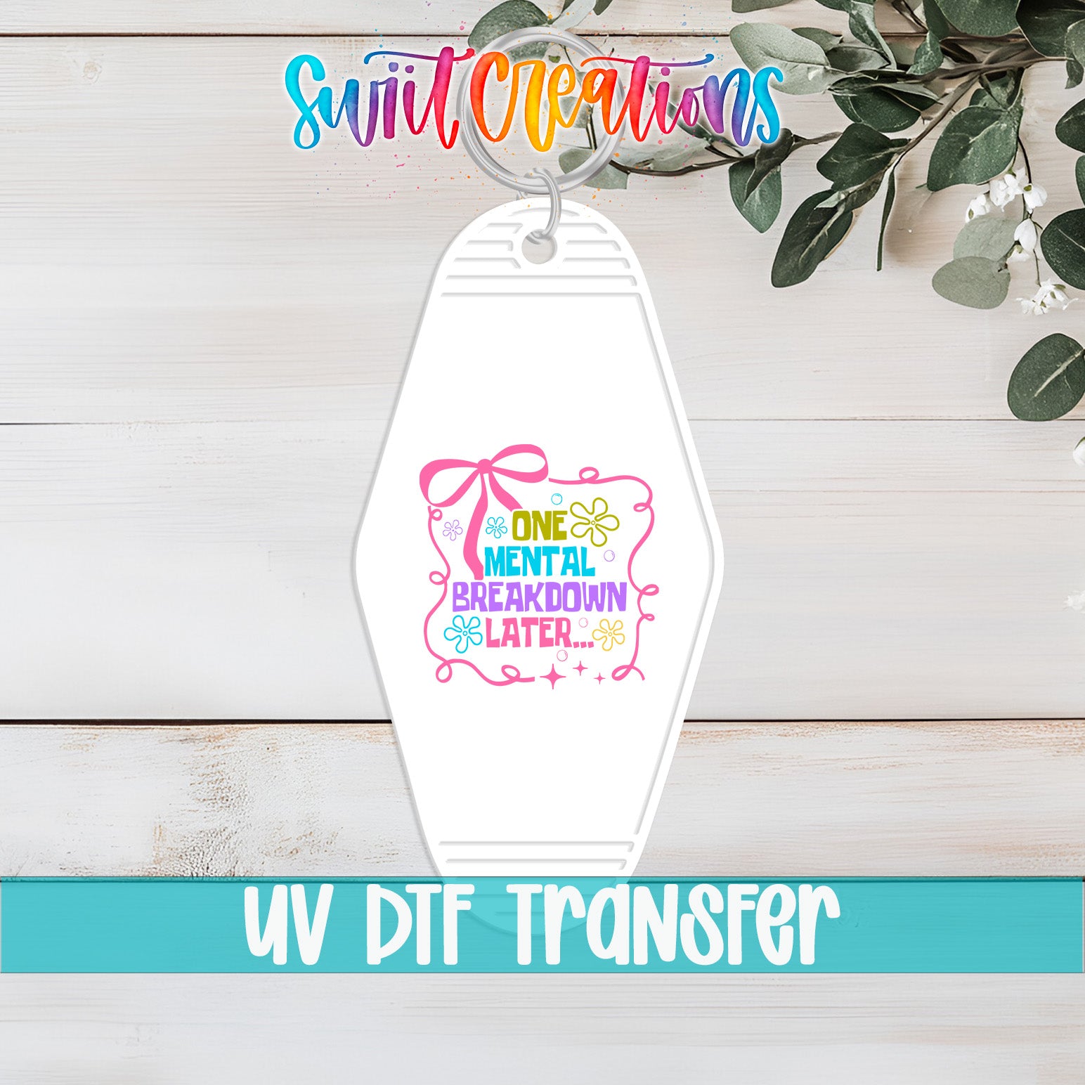 A white keychain with a colorful graphic design and text, placed on a wooden surface with greenery in the background.