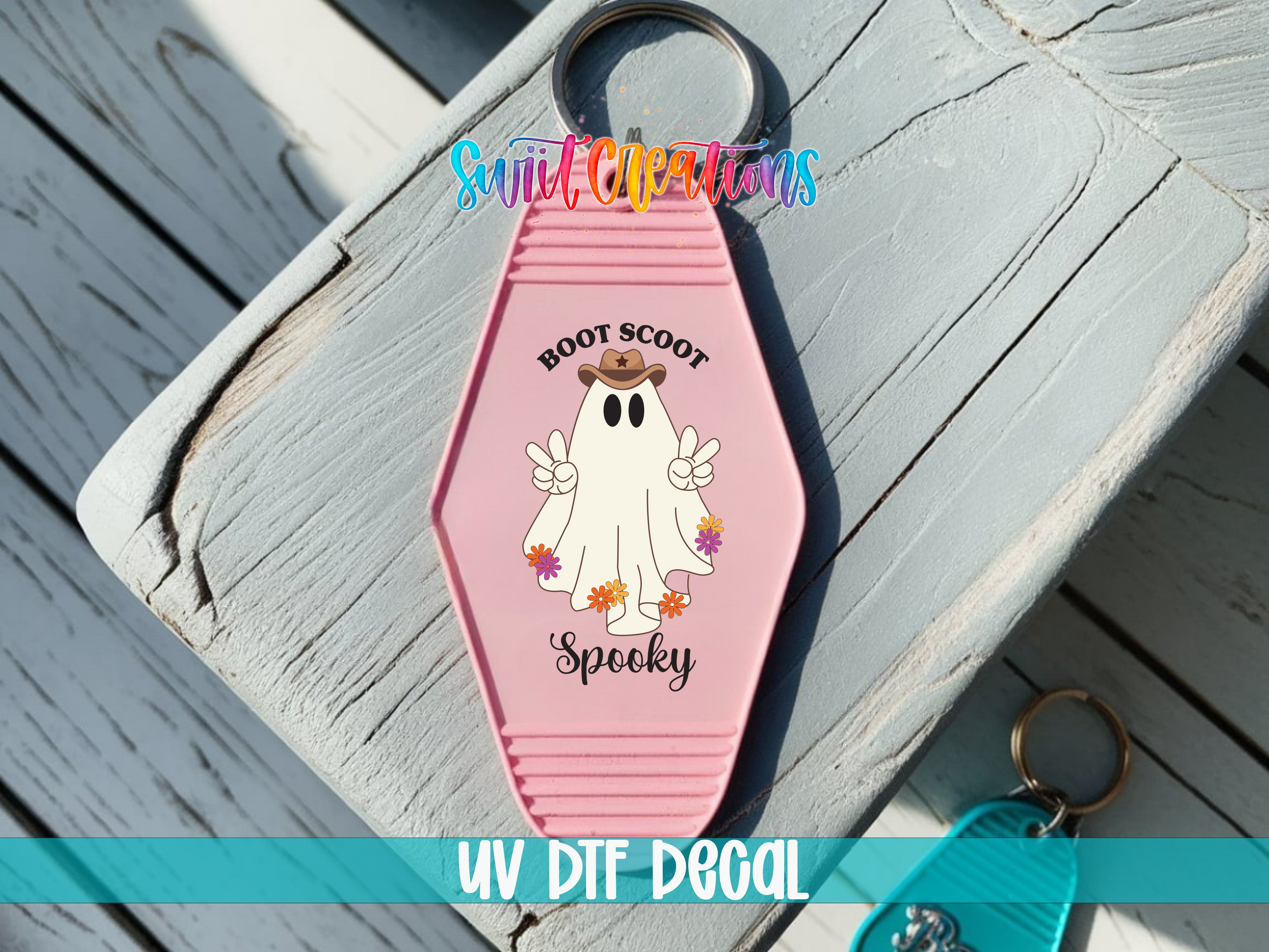 A pink keychain with a cartoon ghost and the text "BOOT SCOOT Spooky" printed on it, placed on a wooden surface.