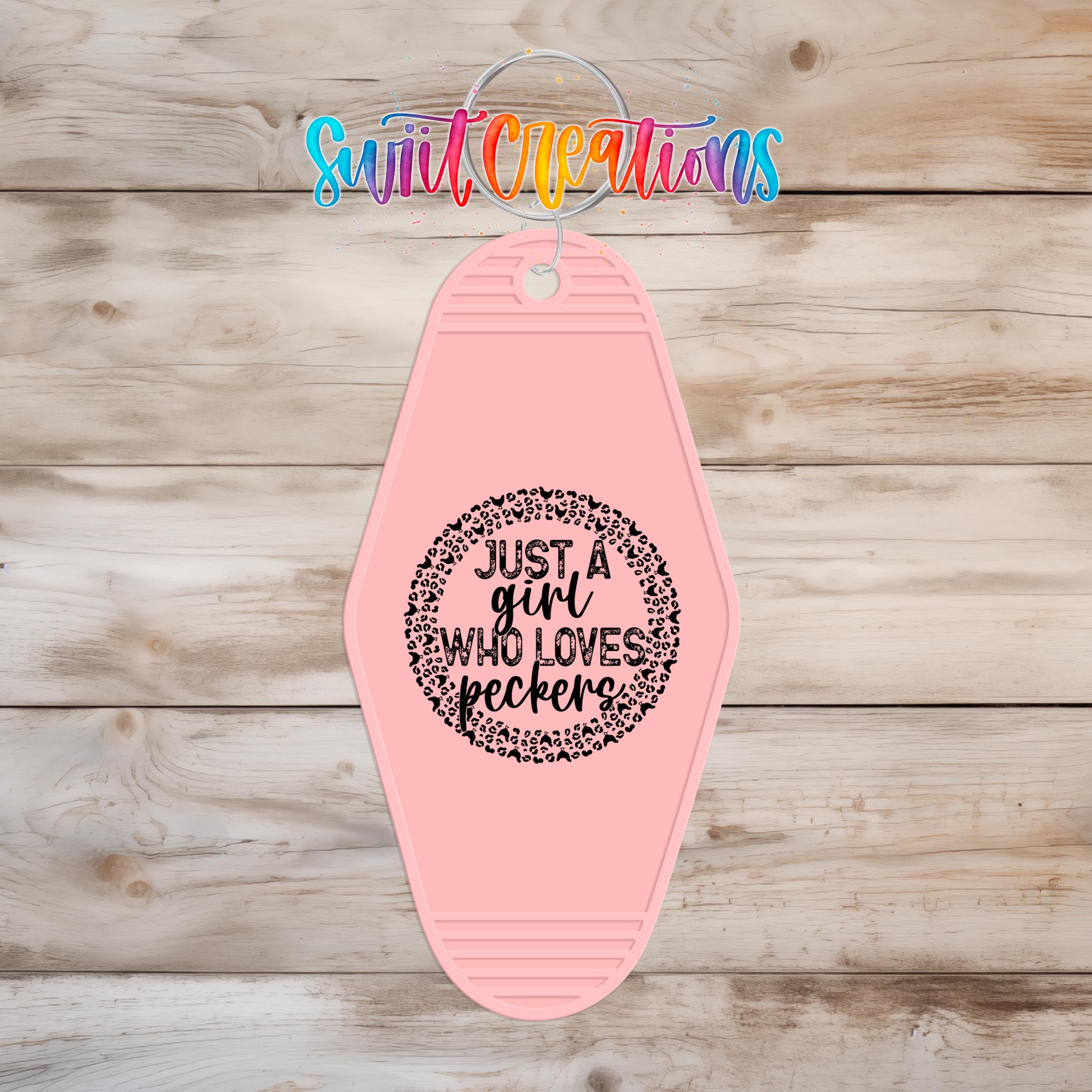 A pink keychain with a black text design that says "Just a girl who loves peckers" on it, hanging on a wooden surface.