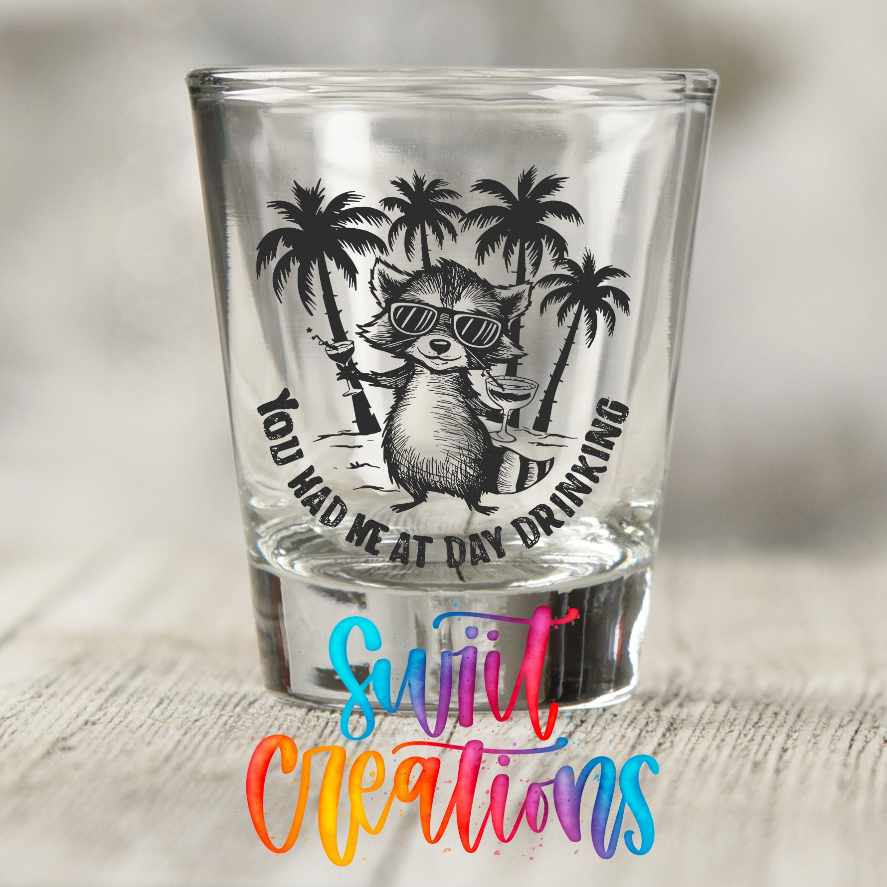 A shot glass with a raccoon wearing sunglasses and holding a drink, surrounded by palm trees, with the text "You had me at day drinking" and "Suit Creations" written below.