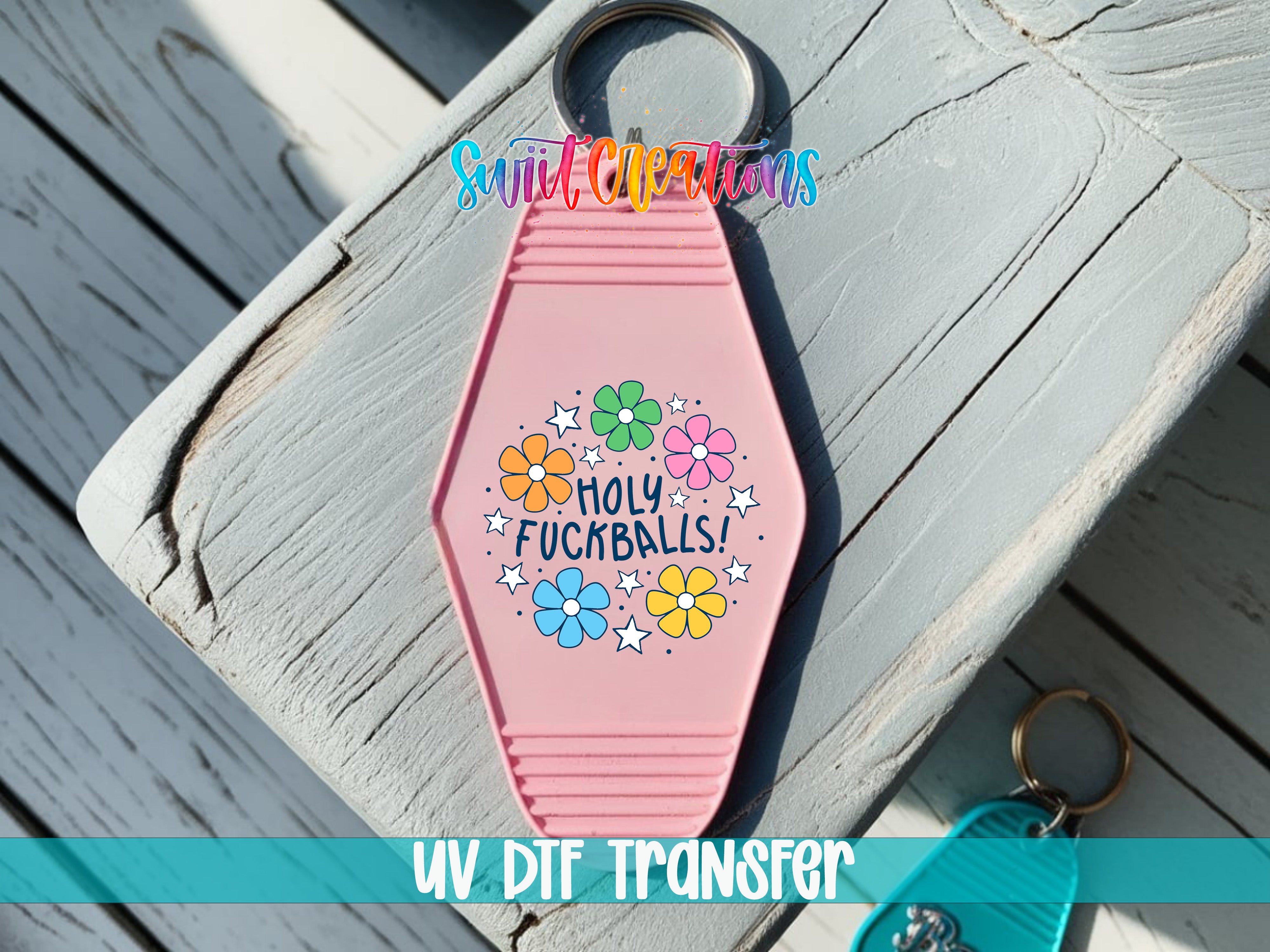 A pink keychain with a floral design and the text "HOLY FUCKBALLS!" printed on it, placed on a wooden surface.