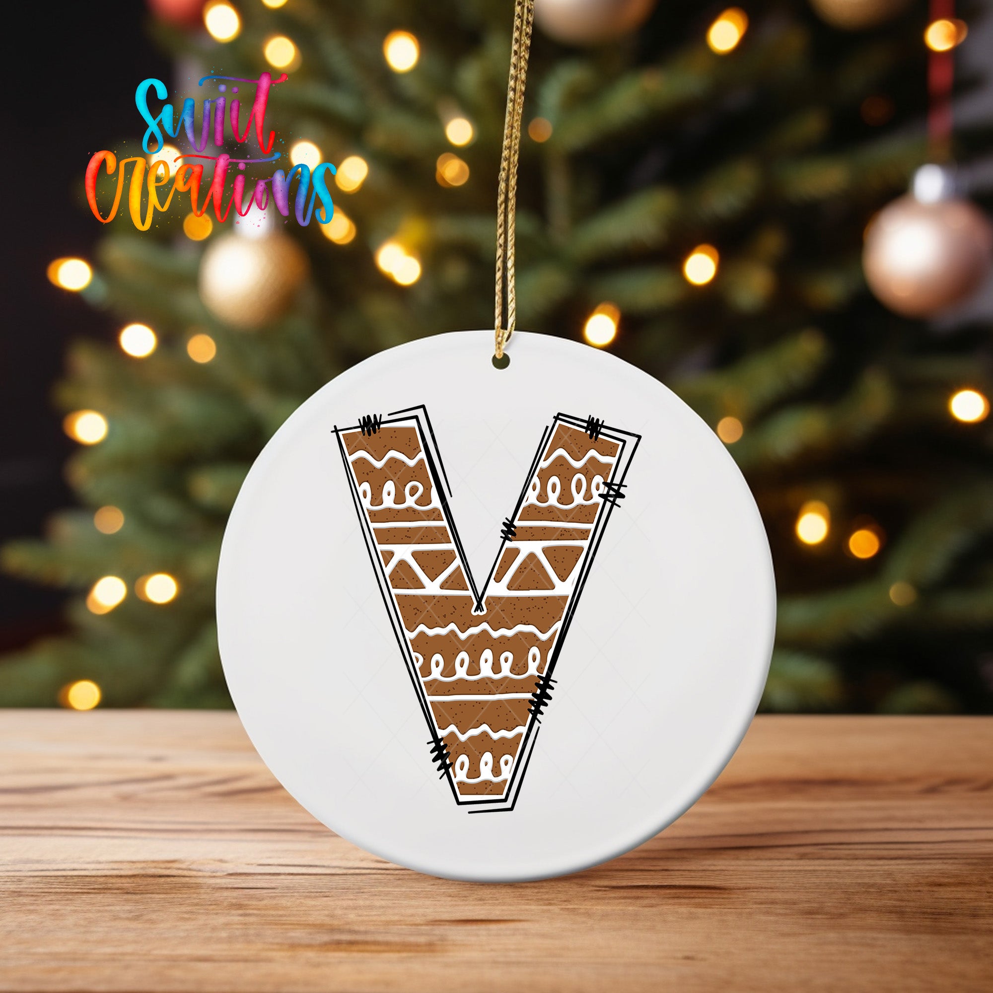 A white circular ornament with a brown design resembling a letter "V" hangs on a wooden surface, with a blurred background of a Christmas tree and twinkling lights.