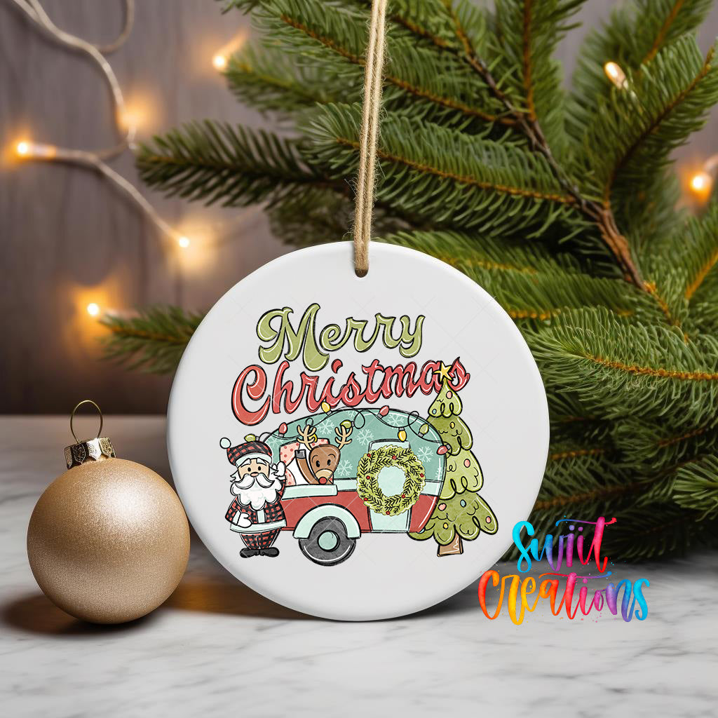 A white ceramic ornament with a Christmas-themed design, featuring Santa Claus, a camper, and reindeer, is displayed on a table next to a gold ornament and a Christmas tree.