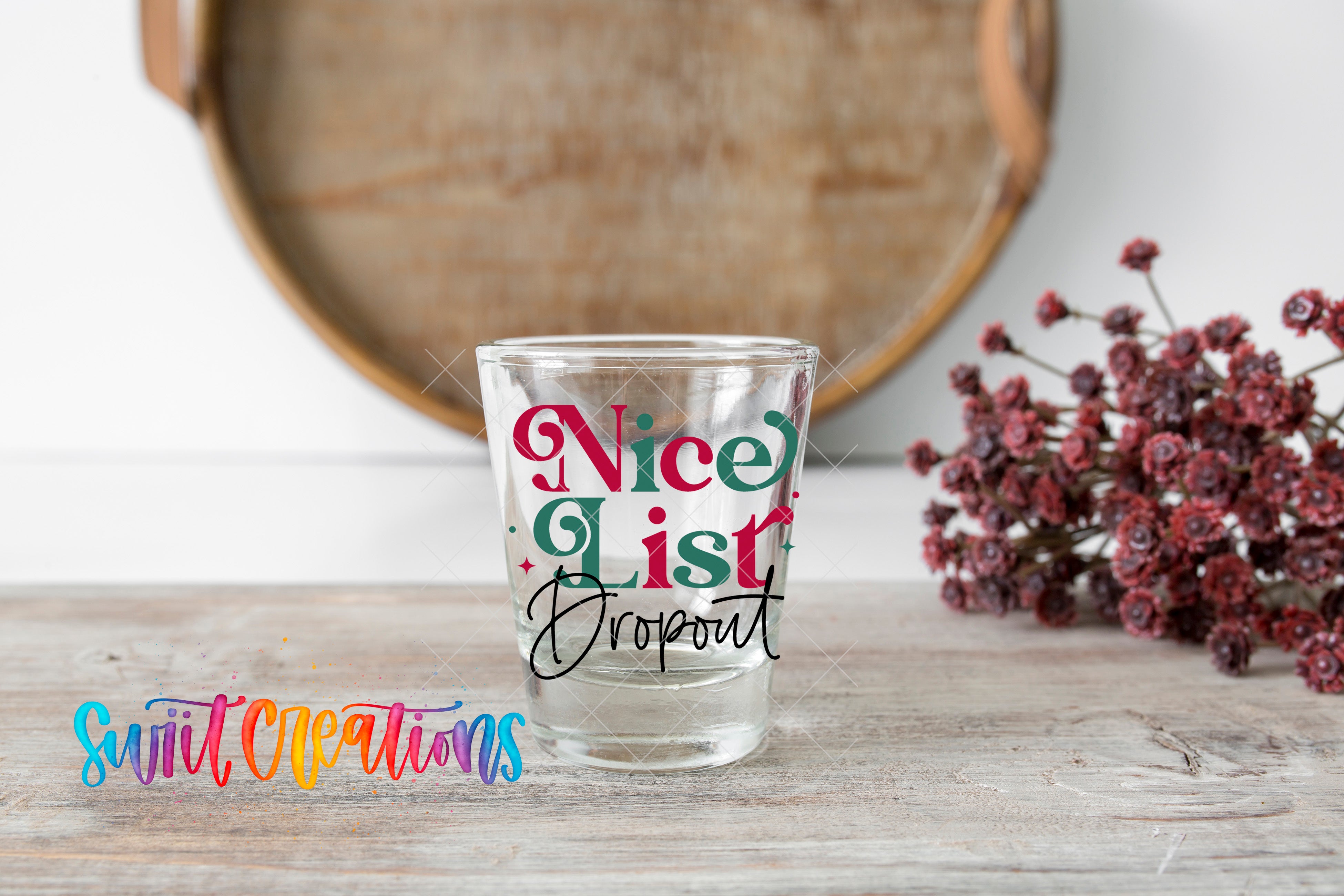 A clear shot glass with the text " Nice List Dropout" printed on it, placed on a wooden surface next to a bunch of red flowers.