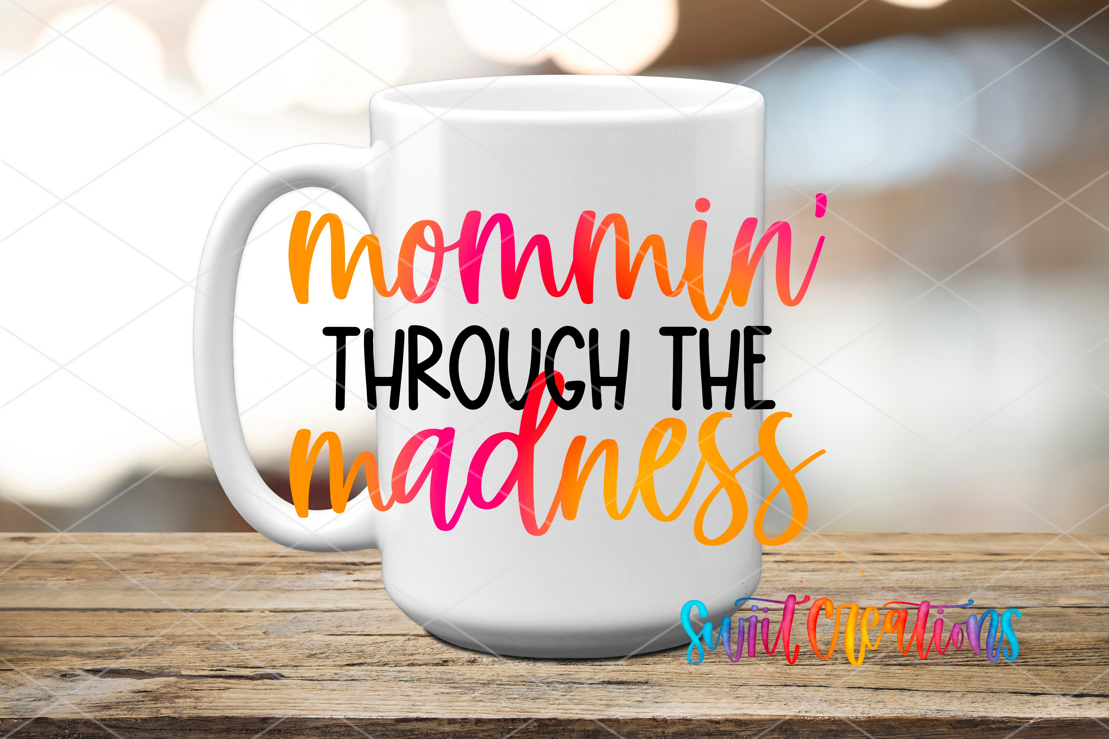A white mug with colorful text that says "mommini THROUGH THE MADNESS" on it, placed on a wooden surface.