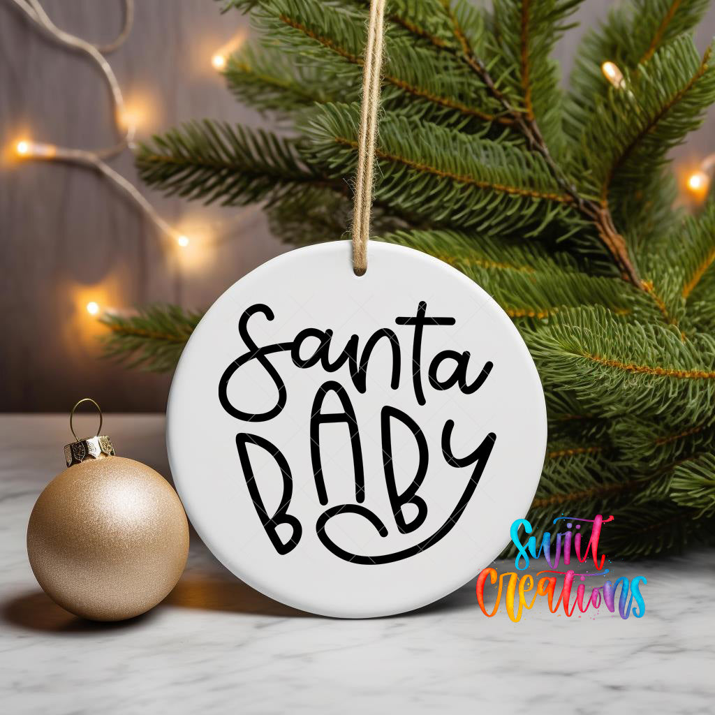 A white ornament with the text "Santa Baby" written on it, hanging from a string, is placed next to a gold ornament and a pine tree with twinkling lights in the background.