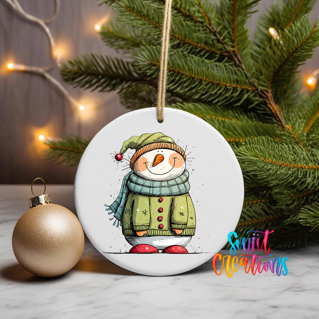 A white ceramic ornament featuring a snowman wearing a green sweater and red shoes, with a Christmas tree and twinkling lights in the background.