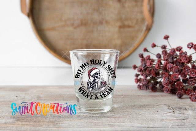A shot glass with a graphic design featuring a skeleton wearing a Santa hat and the text "HO HO HOLY SHIT WHAT A YEAR" is placed on a wooden surface.