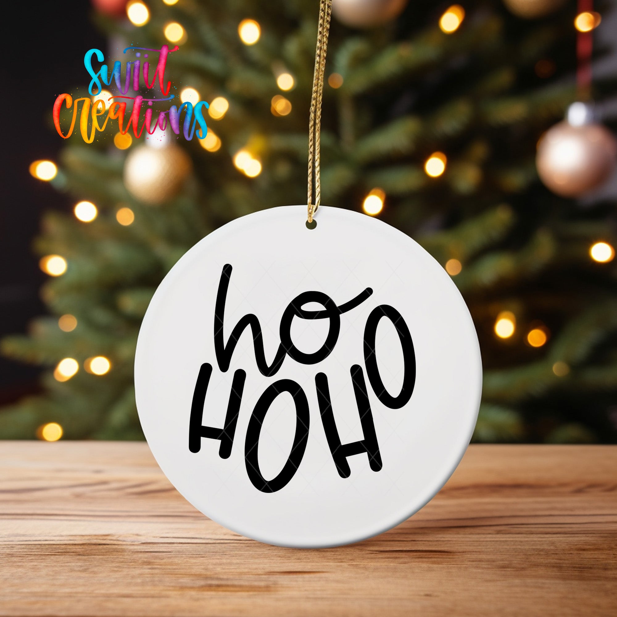 A white ornament with the text "ho ho" hanging from a string, placed on a wooden surface in front of a blurred Christmas tree with colorful lights.