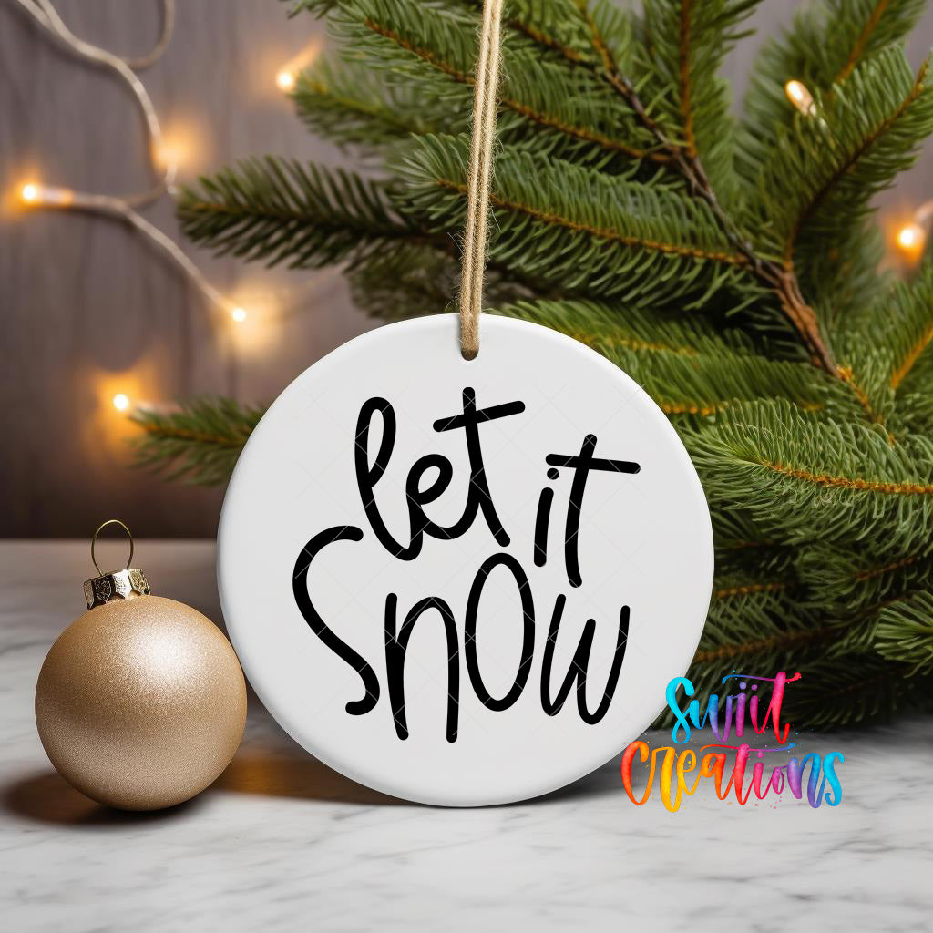 A white ornament with the text "let it snow" written on it, hanging from a string, is placed next to a gold ornament and a pine tree with twinkling lights in the background.