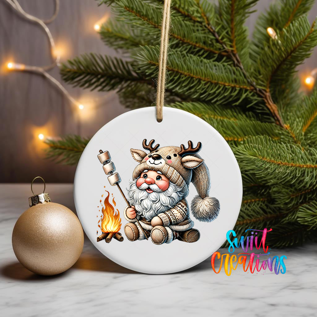 A white ornament featuring a cartoon Santa Claus holding a marshmallow over a campfire, with a Christmas tree and twinkling lights in the background.