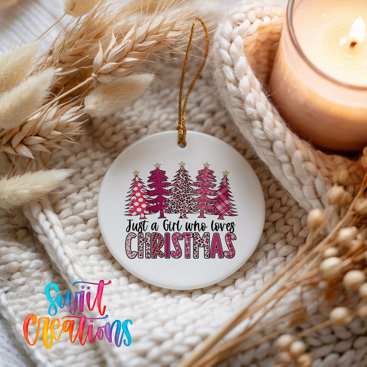 A white Christmas ornament with a design of pink and white Christmas trees, and the text "Just a girl who loves Christmas" is displayed on a white knitted blanket.